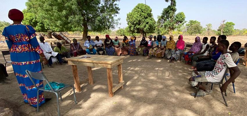 A group of people sitting outside in a semicircle in plastic chairs facing a standing person who appears to be speaking to them.