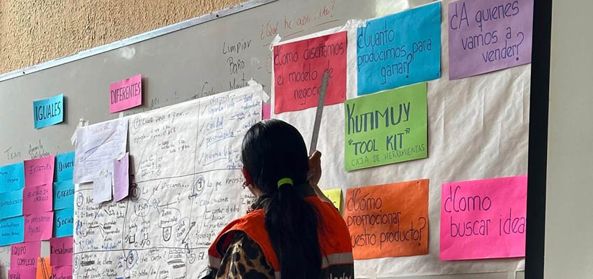 A women with a dark pony tail gesturing towards a wall with post-its and signs.