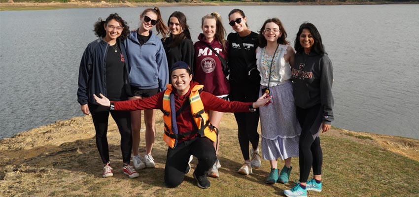 Seven young women standing in front of a stillbody of water and a young man kneeling in front with his arms open wide.