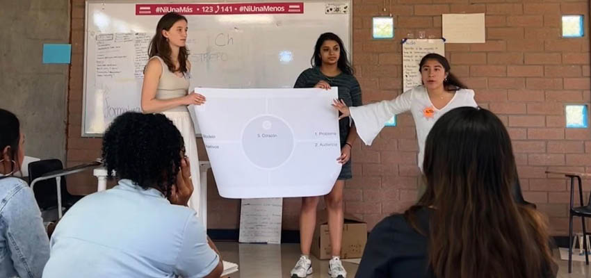 Three women standing showing a sketch on a large piece of paper to three seated people in front of them.