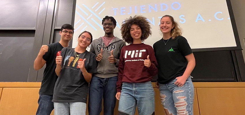 Five standing, smiling college-age people - some giving the thumbs-up sign