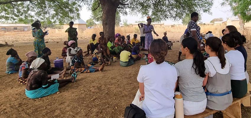 Group of twenty or more women meeting under a tree while a group of 6 young women on a bench look on.