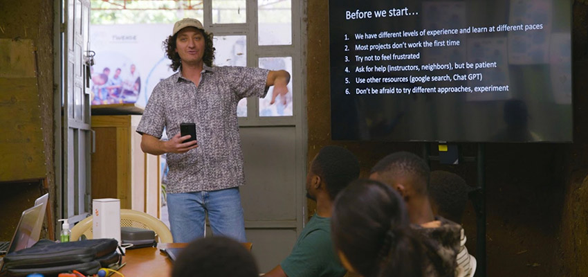 A man in a cap standing next to a large screen with text on it, in front of small group of people seated at a table.