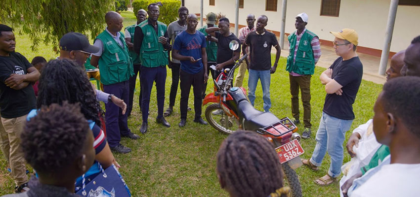 A group of 12+ people standing outside on a patch of grass around a motorbike.