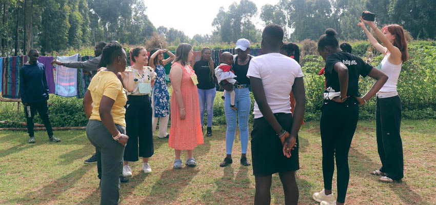 Ten or so people, one holding a baby, standing around outside engaged in an activity.