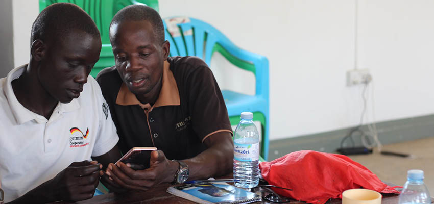 Two men sitting side by side at a table, looking at a cell phone together.