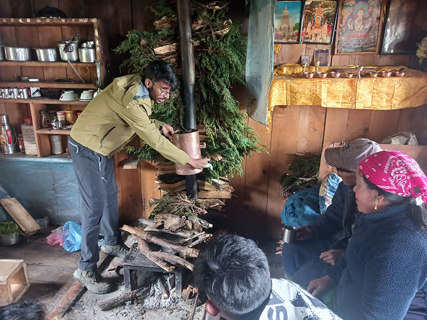 Young bearded man standing by the narrow chimney of an interior cookstove. Three people sit on the floor at right near the stove.