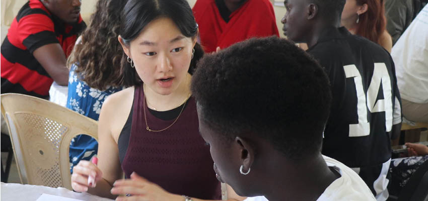 A young woman seated at table speaking to another person who is facing her, also at the table. There are a few people in the background, also seated at table.