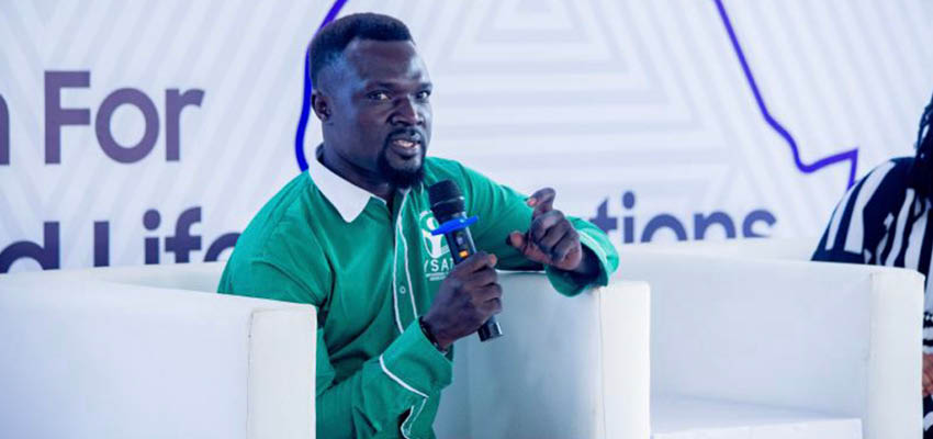 Man sitting in white chair holding a microphone, speaking, and pointing.